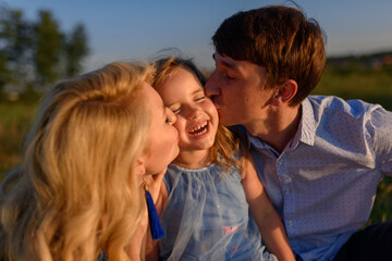 Dad mom and daughter are sitting together on the grass. Parents kiss their daughter on the cheeks.