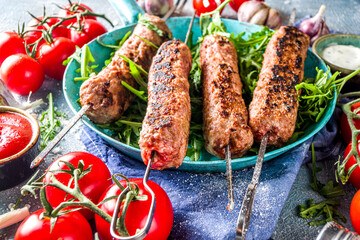 Shish kebab on skewers, from minced beef meat. Lula kebab, traditional Caucasian recipe. On blue concrete background, with sauces,  tomatoes and veggie salad top view copy space