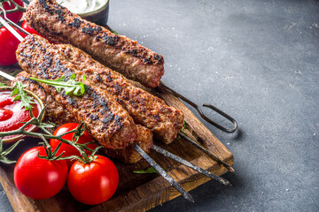 Shish kebab on skewers, from minced beef meat. Lula kebab, traditional Caucasian recipe. On black concrete background, with sauces, tomatoes and veggie salad top view copy space