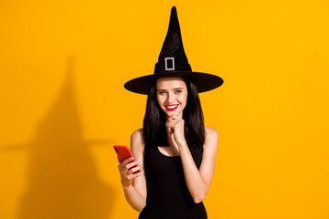 Photo of cute lovely young magician lady hand chin hold telephone beaming smiling post picture event social network wear black wizard headwear dress isolated bright yellow color background