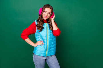 Photo of pretty cute young lady hands hip beaming shiny smiling warm cafe winter weather wear pink ear warmers jeans blue zipped vest red sweater isolated green color background