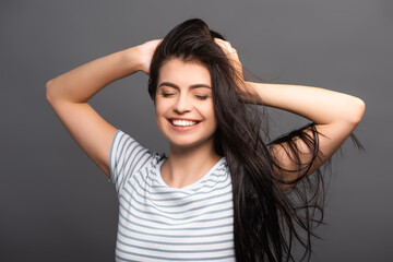 Fototapeta premium brunette woman with closed eyes smiling and touching hair isolated on black