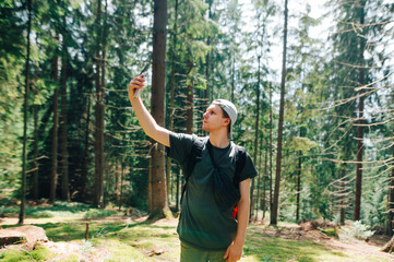 Young male tourist looking for a cellular network on a smartphone in a beautiful forest, lifted a phone up and looks at the screen with a serious face. Guy uses the Internet on a smartphone on a hike