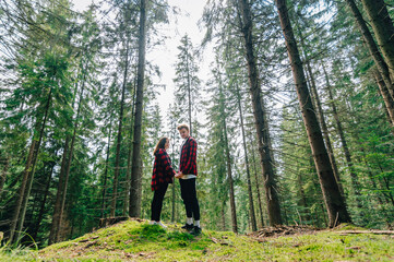 Naklejka na ściany i meble Young couple man and woman in red shirts stand in the woods on the moss, holding hands and posing for camera. Couple of hipsters on a hike in the woods holding hands on a background of beautiful trees