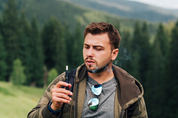 Adult male tourist in casual clothes smokes an electronic cigarette on a background of a mountain...