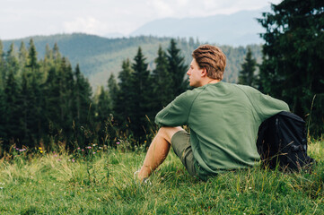 Back view a man with a backpack sits on a mountain plain among forest trees, looking away. Hipster tourist resting sitting on a meadow in the mountains, looking away