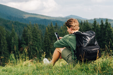Naklejka na ściany i meble Young man with a large black briefcase sits on the soft grass, in a mountain glade, checking phone.Hiker uses a smartphone in mountains sitting on a meadow on the background of a beautiful landscape