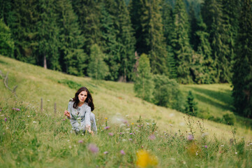 Fototapeta na wymiar Positive girl in a blue dress sits on the grass in the mountains and collects flowers, looks at camera with a smile on his face. Pretty lady in a dress collects grass sitting in the grass on meadow
