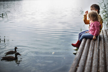 Brother and sister sitting on the river pier and hugging and look at ducks in water. friendship...