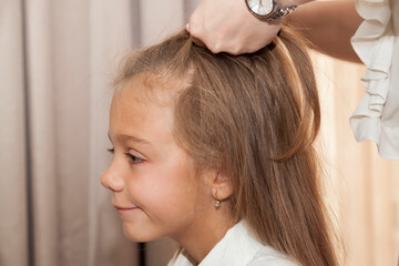 Fototapeta premium Close up photo of happy smiling schoolgirl with long hair and her mother who is combing her hair at home, they are dressed in white blouses, lifestyle people concept