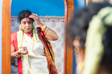 Married Indian Bengali brunette woman wearing traditional red and white sari is looking her face in...