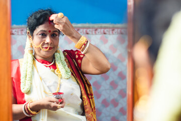 Married Indian Bengali brunette woman wearing traditional red and white sari is looking her face in...