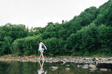 Naklejka premium Stylish lady in casual clothes and a backpack walks on a mountain river, walks on rocks. Tourism in the mountains