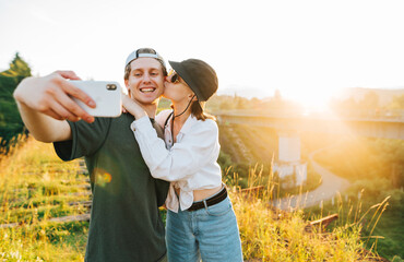 Cute young couple of tourists taking a selfie while walking on the old bridge, a young man looking at the camera of a smartphone, a girl kisses a guy on the background of a bitter landscape at sunset
