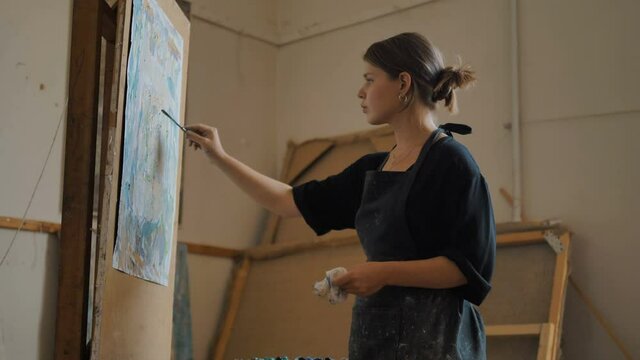 concentrated female artist in black apron applies paint on canvas with small spatula drawing picture in spacious art studio
