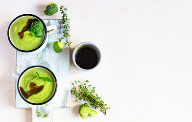Two enamel mugs with healthy vegan broccoli soup with spicy oil and aromatic herbs over light stone background. Diet detox food concept. Top view, copy space.