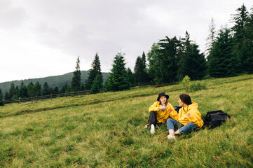 Fototapeta na wymiar Two girls tourists in yellow raincoats sitting on a meadow in the mountains against the backdrop of a beautiful landscape, relaxing and talking. Hikers rest on a meadow.
