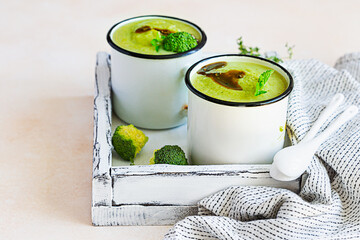 Two enamel mugs with healthy vegan broccoli soup with spicy oil and aromatic herbs over light stone background. Diet detox food concept.