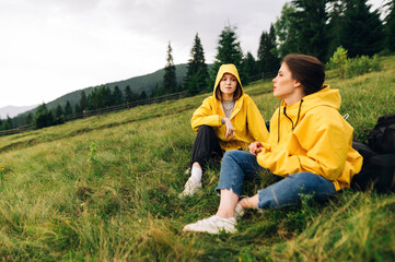 Fototapeta premium Two attractive women in yellow raincoats sitting on a meadow in the mountains against the backdrop of a beautiful landscape and talking.Tourists resting on a hike sitting on a field mountain landscape