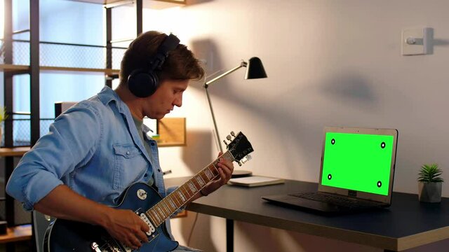 leisure, music and people concept - young man or musician in headphones with green chroma key screen on laptop computer playing bass guitar sitting at table at home