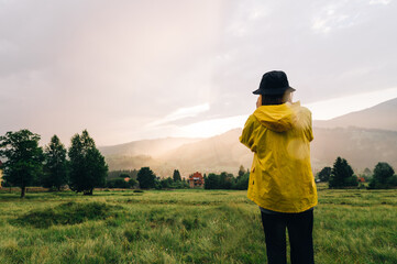 A person in a yellow raincoat stands on a meadow in the mountains on a background of sunset and takes a photo, view from the back. Girl in panama and raincoat on the background of a mountain village