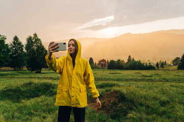 Attractive girl in a yellow raincoat takes a selfie on a background of beautiful sunset in the mountains. Woman tourist makes a photo in the rain at sunrise