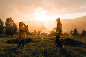 Two hikers in yellow raincoats standing on a meadow in the mountains against the backdrop of an unreal sunset and taking photos on a smartphone. Two female hikers in the rain at sunset
