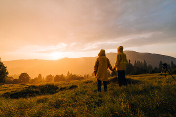Fototapeta na wymiar People in yellow raincoats stand on a meadow in the mountains and watch the beautiful sunset in the rain and shoot with a camera on a tripod. Background. Hikers in the rain at sunset on a hike