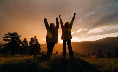 Two hikers in yellow raincoats stand on a meadow on a background of sunset in the mountains with raised arms. Silhouette of joyful tourists on a background of sunset in the rain