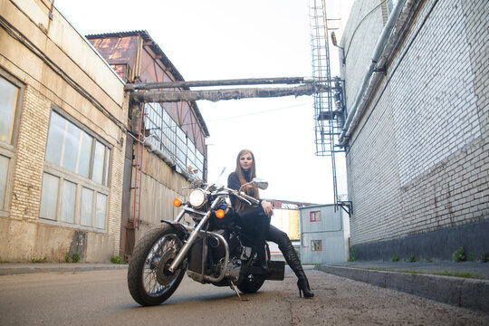 A beautiful slender girl biker sits on a motorcycle against the backdrop of an industrial city building.