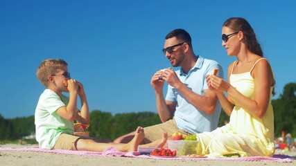 family, leisure and people concept - happy mother, father and little son having picnic on summer beach and eating sandwiches