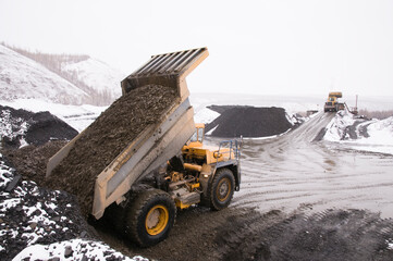 Winter in a mountainous wooded area. Earthworks - extraction of minerals. A dump truck unloads mountain soil, a wheel loader is working in the background.