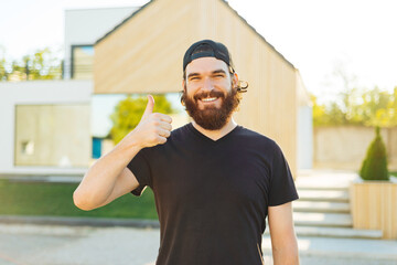 Fototapeta premium Happy cheerful young man showing thumbs up and standing in front of his modern house