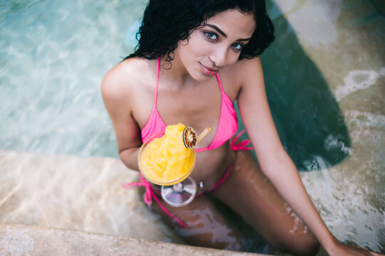 From above picture of brunette female in pink swimsuit in pool holding fresh beverage for refreshment, attractive woman with slim figure enjoying relaxing on villa resort during holiday vacations