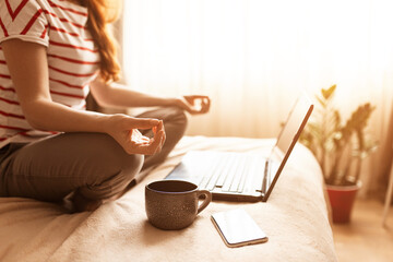 Young calm woman sits in staff easy lotus pose near laptop at home interior, meditating alone on...