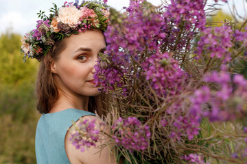 Fototapeta premium Slavic beauty with a floral wreath on her head and a bouquet of wildflowers. Ancient pagan origin celebration concept. Summer solstice day. Mid summer. Ancient rituals.