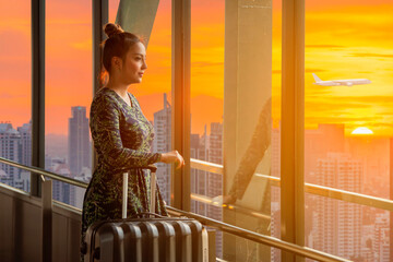 Smart businesswoman pulling a suitcase in a big city looking out the window of high rise skyscraper building and Airplane , Beautiful sunset on background.