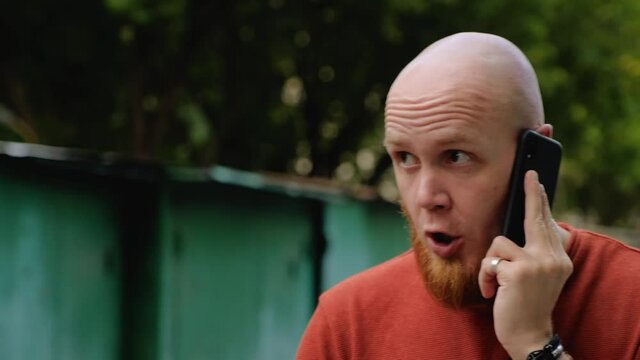 A young bald guy with a red beard in a dark orange sweater on the street speaks seriously and shouts on the phone. Conversation in a raised voice. Modern gadget. Dialogue. Emotions of the face.