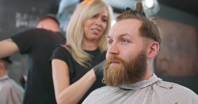 Closeup face of young bearded man, who is sitting on the barber's chair, facing the mirror, and female barber clippers his hair with hair clipper and comb, and talks to him. Another barber with client