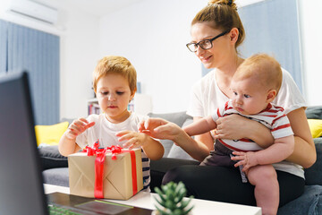 Single Mother with two small kids with wrapped gift box at home in front of laptop - Caucasian woman sitting on sofa sharing present with son - celebration new normal covid-19 social distance concept