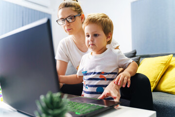 Small boy standing by his mother in front of laptop at home - Little kid using computer to watch online video or make a call