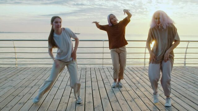 Dancing group of young talented freak women performing freestyle hip-hop moves. Girls enjoying modern dance expression. Outdoor training near sea or ocean during sunset.