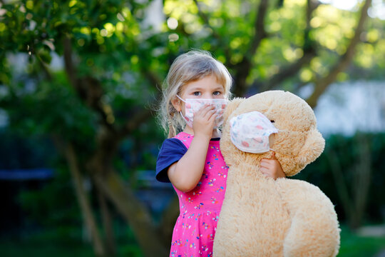 Little toddler girl in medical mask as protection against pandemic coronavirus quarantine disease. Cute child using protective equipment as fight against covid 19 and holding big bear toy with mask.