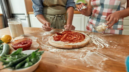 Cropped shot of couple making pizza together. Man in apron adding, applying tomato sauce on the dough while woman helping him in the kitchen. Hobby, lifestyle