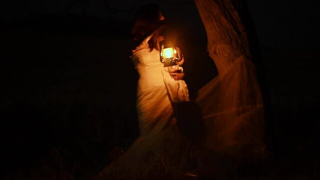 Scary woman with a lantern in night scene - Spooky image of a scary woman with dark eyes and appearance of a witch, in a white dress, holding a lit lantern and a frightening doll, in a dark night