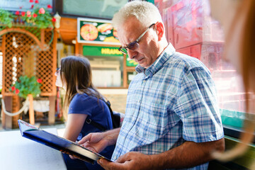 Side view on senior man sitting in restaurant in sunny summer or autumn day checking the menu getting ready to order - real people tourism concept