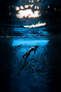 A female freediver with bikini and fins diving in the cave with lots of fish
