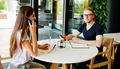 Portrait of handsome hipster guy in spectacles for provide eyes protection looking at camera while planning organisation, Caucasian female using wireless internet connection for call phoning