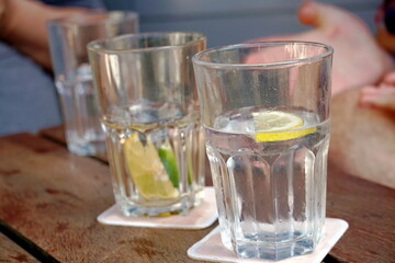 Glasses with water, lemon and lime slices on a table in an outdoor restaurant. The glasses are fogged or steamed on a hot summer day. People sit around and talk, only their hands are visible. 
