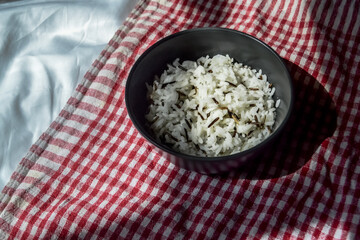 white parboiled black rice in a dark cup on plate on a background of red-white cloth towels under the light of the sun with a shadow on the table